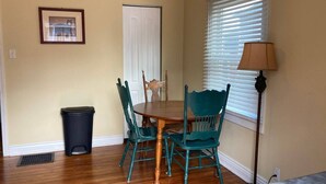 Un comedor con una mesa de madera y sillas color turquesa, un cubo de basura, una puerta y una ventana con persianas.
