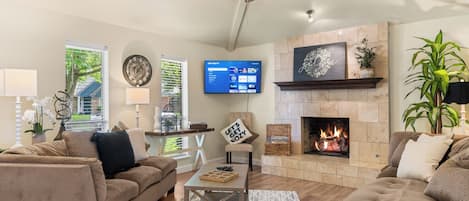 A living room with a fireplace, a ceiling fan, a flat-screen TV, and a large window with a view of greenery.