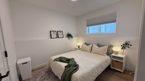A bedroom with a bed, bedside tables, a waste basket, a window, and a plant. - The Soft Fern Retreat | Cozy 2BR suite in Rosewood (Saskatoon)
