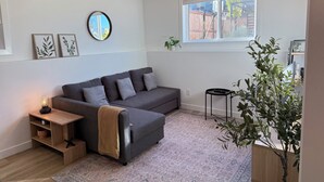 A modern living room with a grey sectional sofa, a small wooden side table, a round mirror, framed botanical prints on the wall, a potted plant, and a window with a view of the outside. - The Soft Fern Retreat | Cozy 2BR suite in Rosewood (Saskatoon)