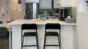 A modern kitchen with white cabinetry, a central island with bar stools, stainless steel appliances, and a microwave. - The Soft Fern Retreat | Cozy 2BR suite in Rosewood (Saskatoon)