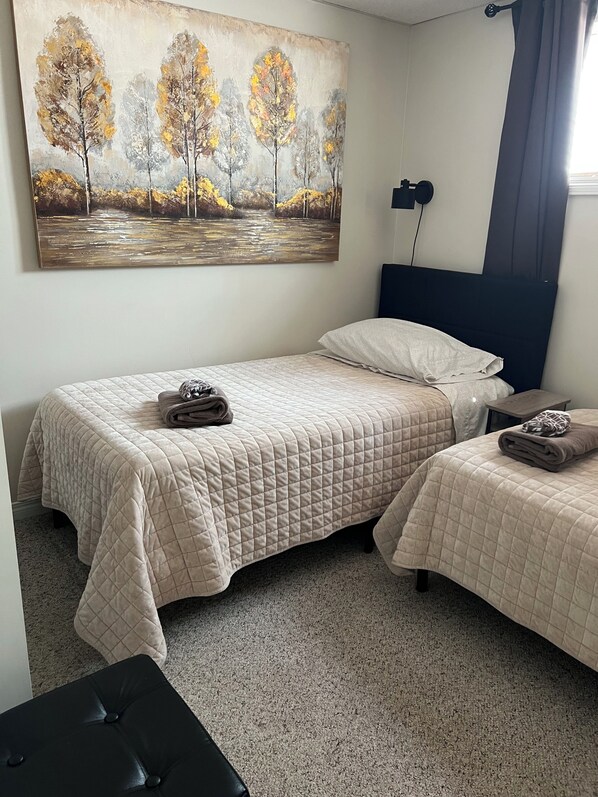 A bedroom with two beds, a painting of trees, a bedside table, and a lamp.