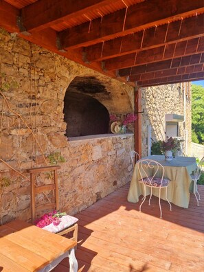 Un four en pierre sous un toit en bois, une table avec des chaises et une petite table sur laquelle est posé un vase de fleurs.