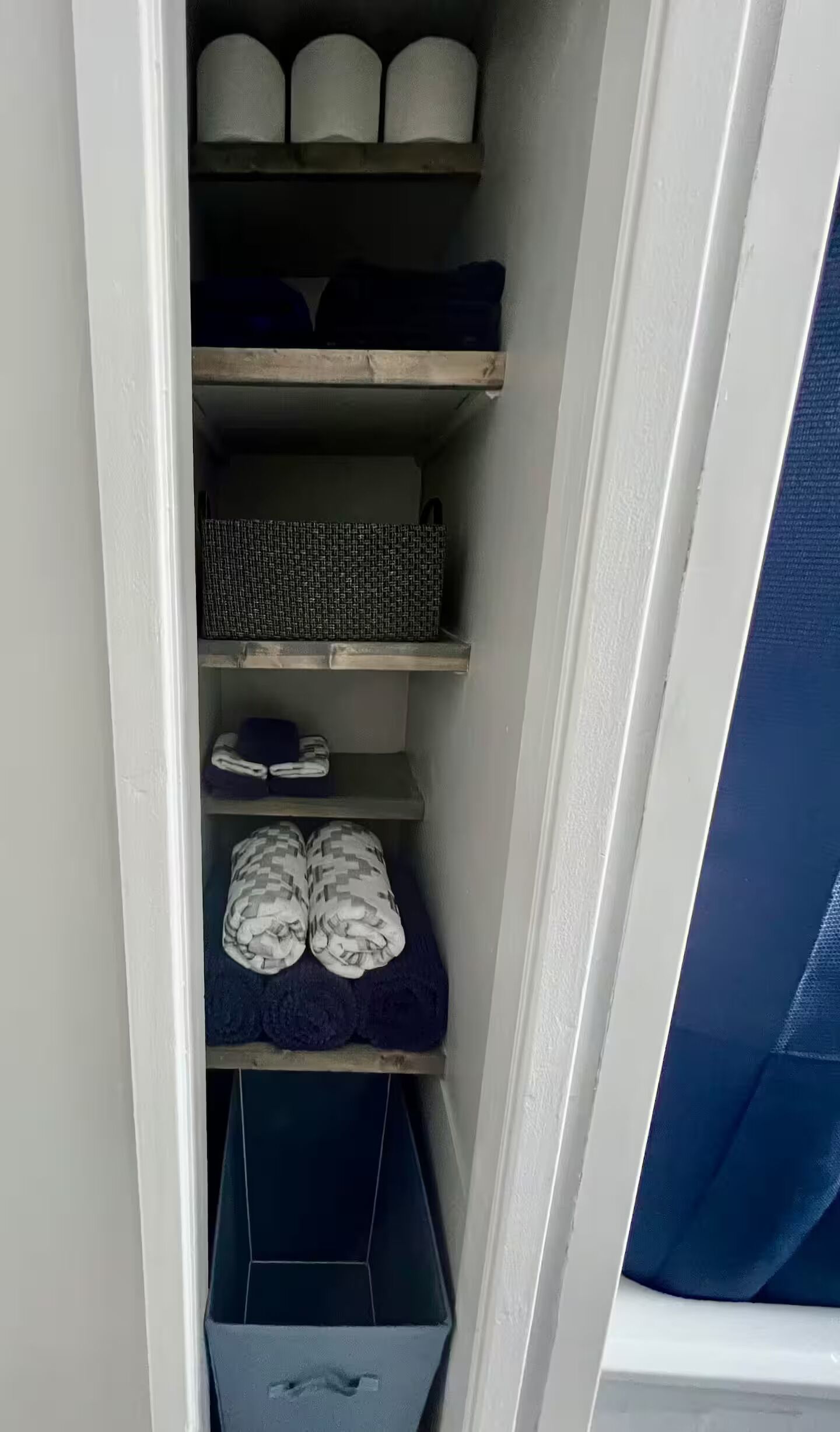 Ein schmaler Stauraum mit Regalen, die verschiedene Gegenstände wie gefaltete Handtücher, einen gewebten Korb und ein Paar Hausschuhe beherbergen.