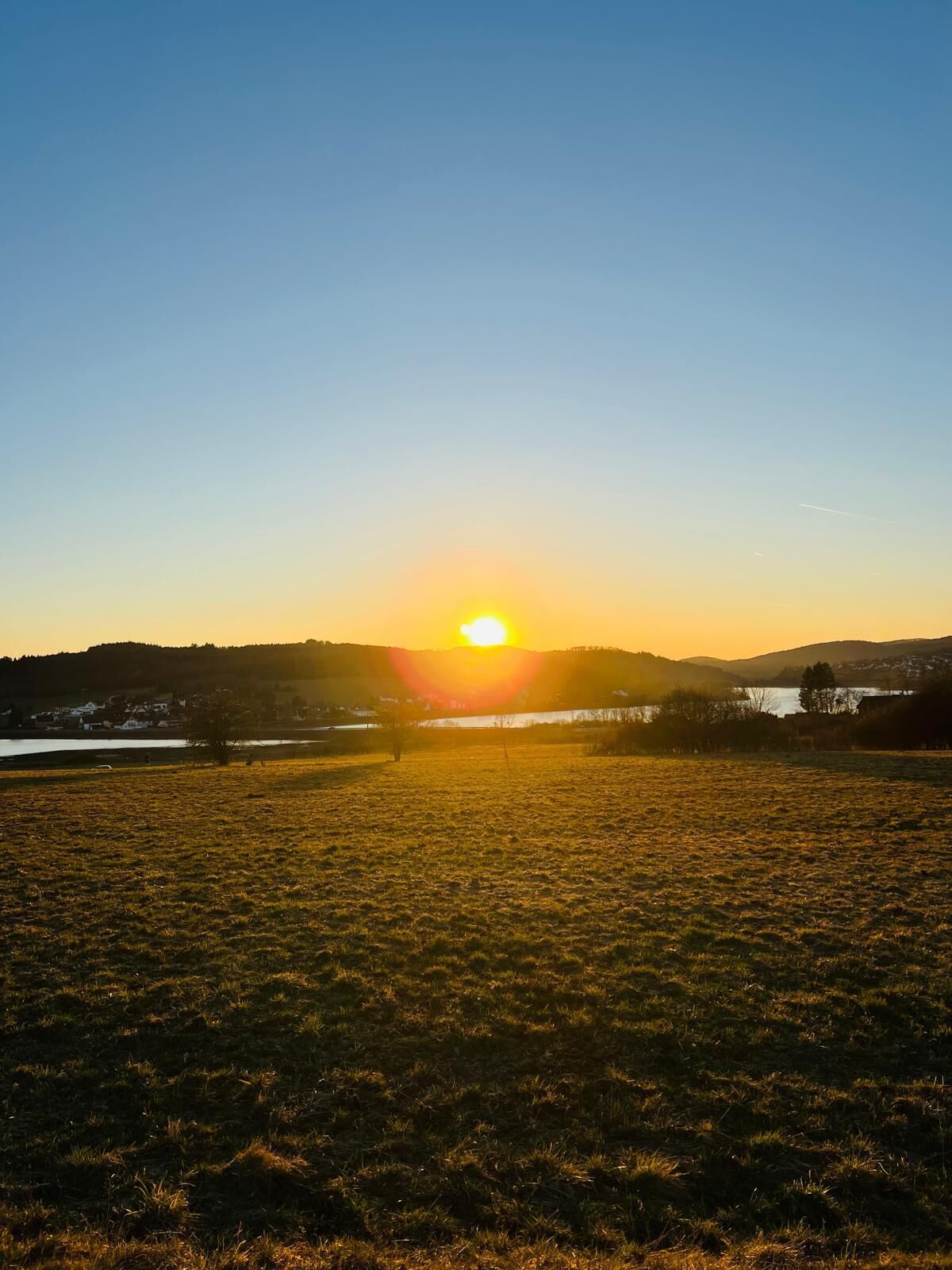 Ein Sonnenuntergang über einem grasbewachsen Feld mit einem See und Hügeln im Hintergrund.