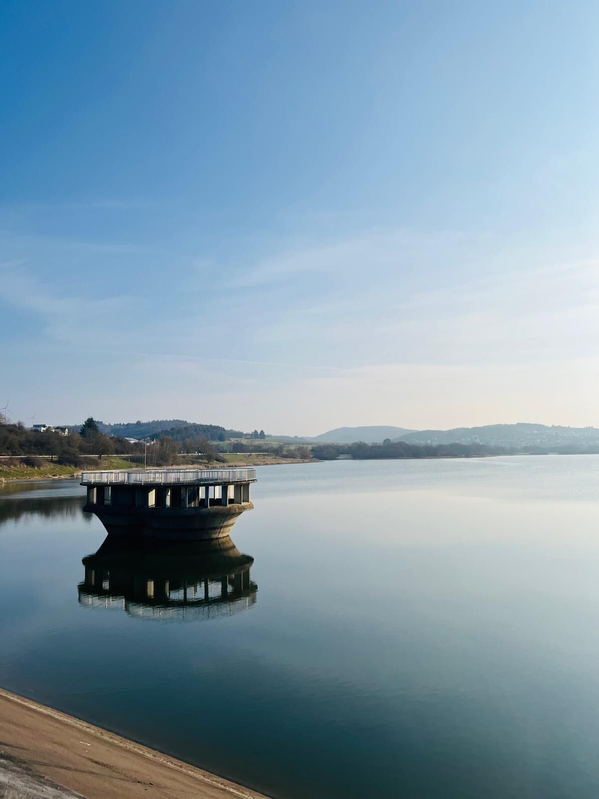 Ein ruhiger See mit einer Betonkonstruktion, die ein Geländer und ein kleines Gebäude auf dem Dach hat, umgeben von einer hügeligen Landschaft.