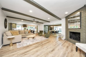A spacious living room with a fireplace, a dining area, and a kitchen visible through an open door. - Trail Manor Oasis | Pool/Htd Spa/Private Arcade+Games+ Cabana (Houston)