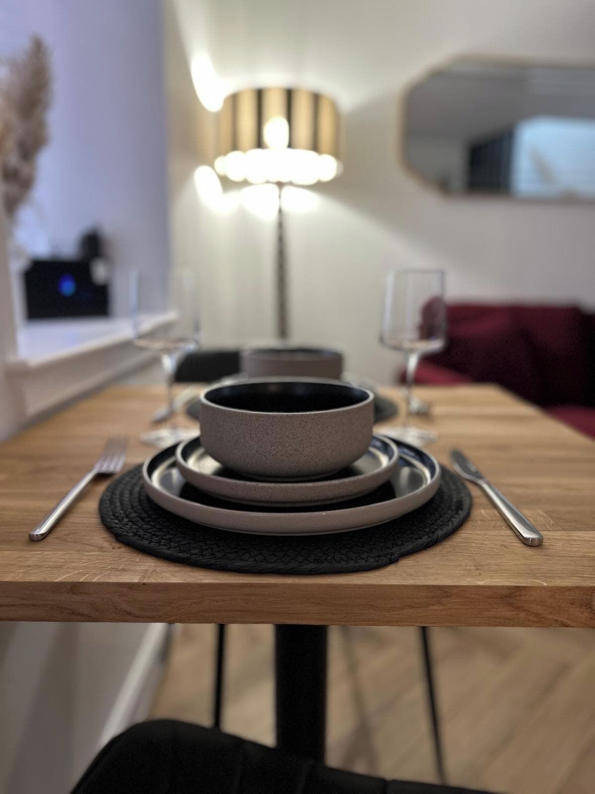 Ein Esstisch mit gestapelten Tellern und Besteck, eine Lampe und ein Sofa im Hintergrund.