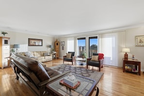 A spacious living room with a leather sofa, a coffee table, and a dining table. There are two armchairs, a side table with a lamp, and a bookshelf. The room has large windows with curtains. - Spectacular View of the Ozarks, Near Golf Course (Holiday Island)