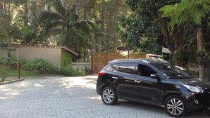 Une voiture noire est garée dans une allée dont la surface est pavée, entourée de verdure et d’une clôture en bois.