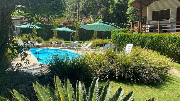 Un espace piscine doté de chaises longues et de parasols, entouré d’une végétation luxuriante et bordé d’un bâtiment avec une terrasse en bois.
