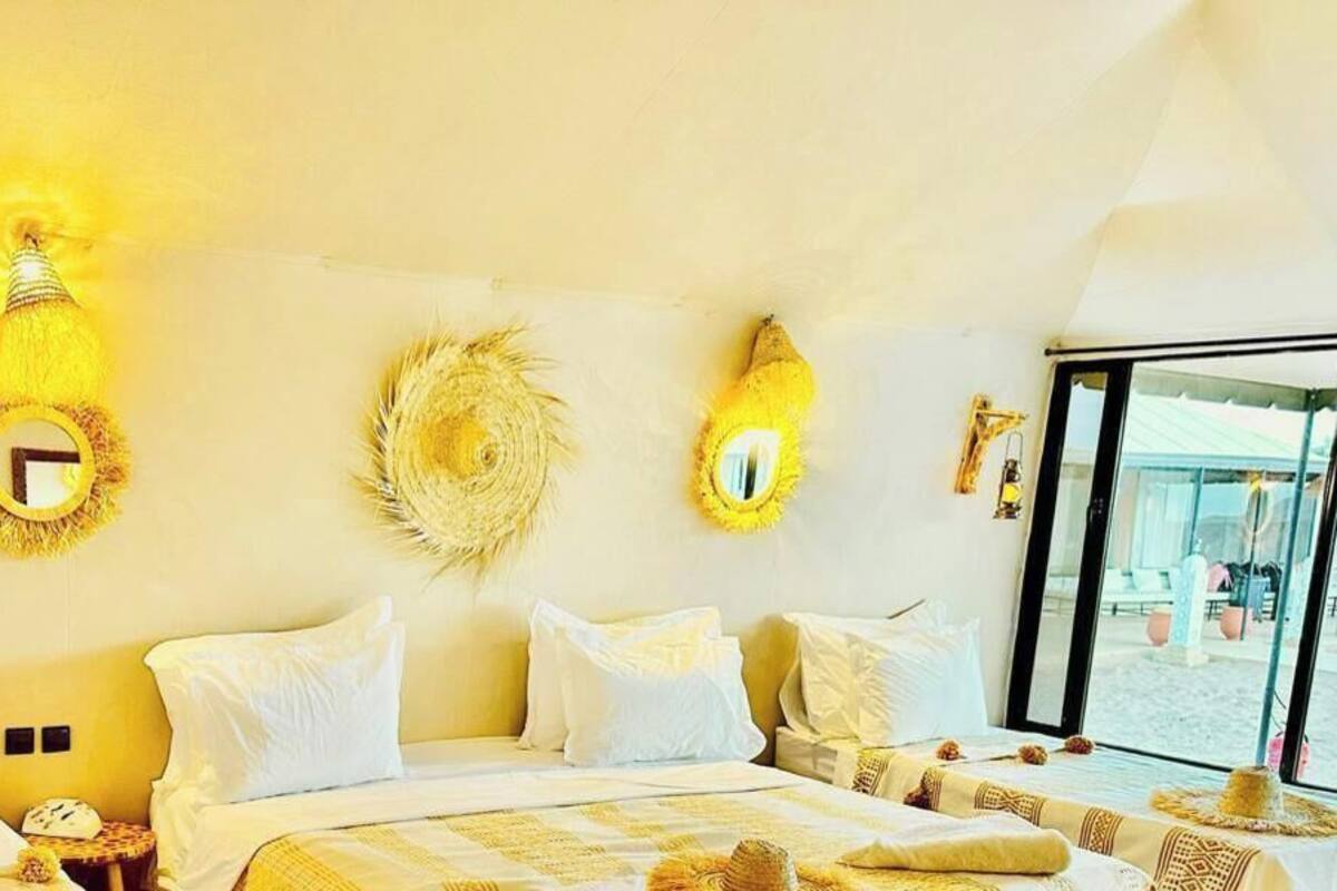 Quarto de hotel com cama grande, várias almofadas, luminárias fixadas na parede e vista através de uma janela ampla.