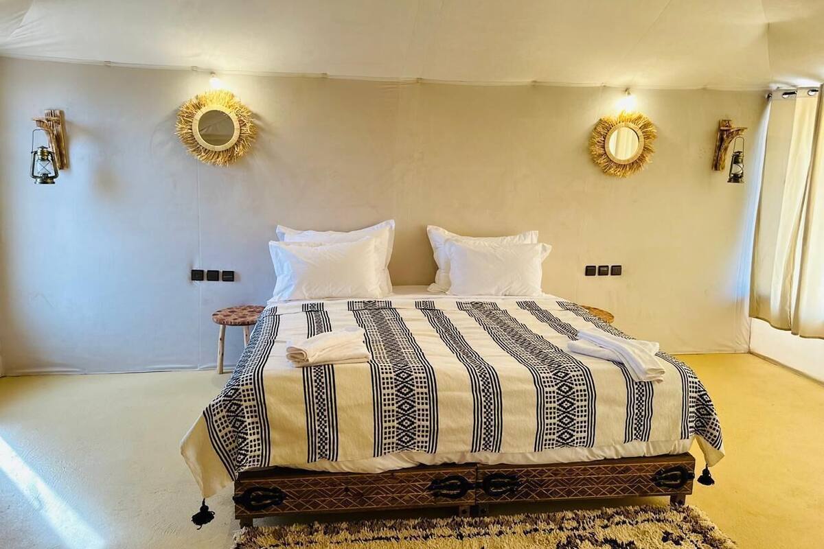 Quarto com uma cama grande, duas arandelas, um espelho redondo e um banquinho pequeno.