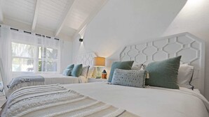 Quarto com duas camas, mesa de cabeceira com abajur e janela com cortinas leves.