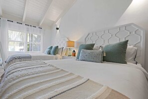 Et soveværelse med to senge, et natbord med lampe og et vindue med let gardin.