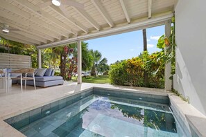 A modern pool area with a clear view of the outdoors, a seating area with a sofa and chairs, and a well-maintained garden.