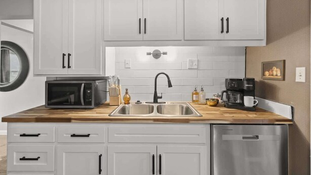 Una cocina moderna con armarios blancos, un lavamanos de acero inoxidable, microondas, cafetera y lavavajillas.