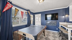 Una habitación con mesa de ping pong, una zona de bar con taburetes y un televisor montado en la pared.