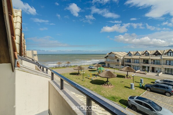 Un hotel frente al mar con balcón con vistas al océano, un parque infantil y varios coches aparcados en la entrada.