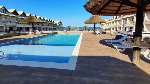 A resort pool area with multiple lounge chairs and umbrellas, surrounded by hotel buildings with thatched roofs. - Houses in a Beachfront Condominium With a Swimming Pool in Itajuba - Mcss (Barra Velha)