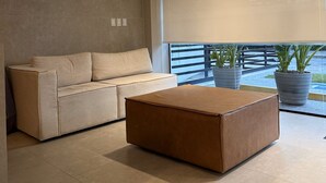 Un salon moderne avec un canapé beige, un pouf marron et de grandes fenêtres équipées de stores.