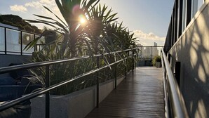 Une terrasse en bois avec des rampes en métal, offrant une vue sur un paysage urbain avec des immeubles et un palmier.