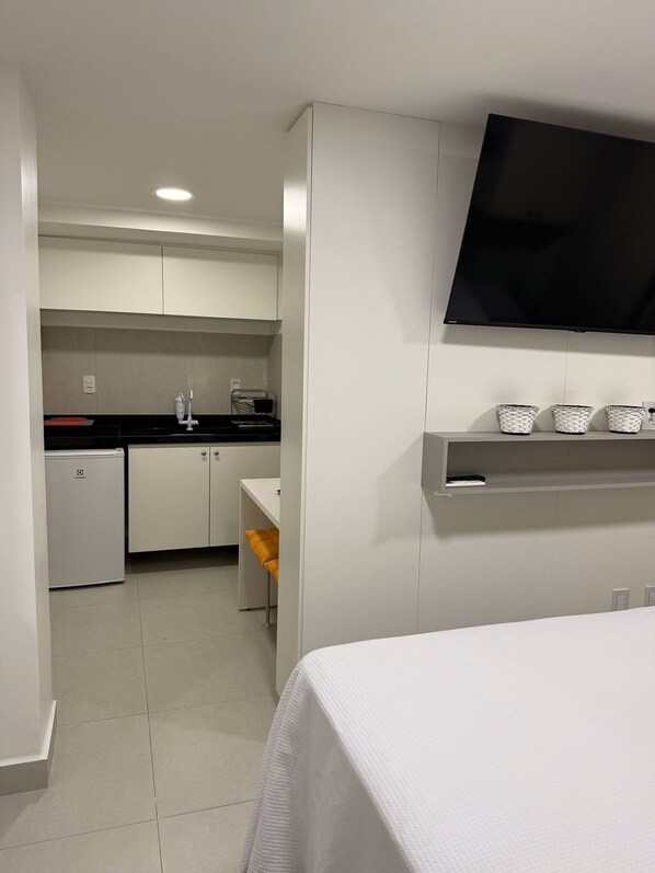 A compact hotel room with a bed, a TV mounted on the wall, a kitchenette with a fridge, and a small table with a chair. - Garden Estudio Cabo Branco 50 m do mar (João Pessoa)