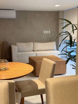 A modern living room with a round wooden table, beige upholstered chairs, a beige sofa, and a large potted plant. - Garden Estudio Cabo Branco 50 m do mar (João Pessoa)