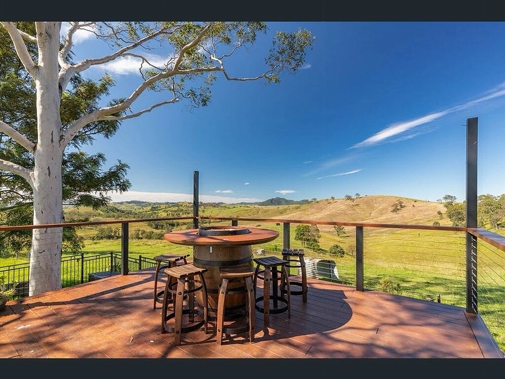 Um deck de madeira com uma mesa redonda e banquinhos, com vista para uma paisagem gramada com colinas.