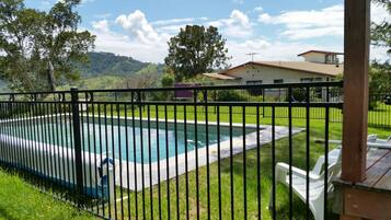 A rectangular swimming pool with a black metal fence, surrounded by a grassy lawn and a wooden deck with white chairs.