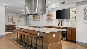 Una cocina moderna con una isla central, taburetes de bar, armarios de madera, una campana extractora de acero inoxidable y un televisor de pantalla plana montado en la pared.