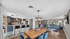 A modern kitchen with bar stools, a dining area with a wooden table and blue chairs, and a living room with a sofa and a coffee table.