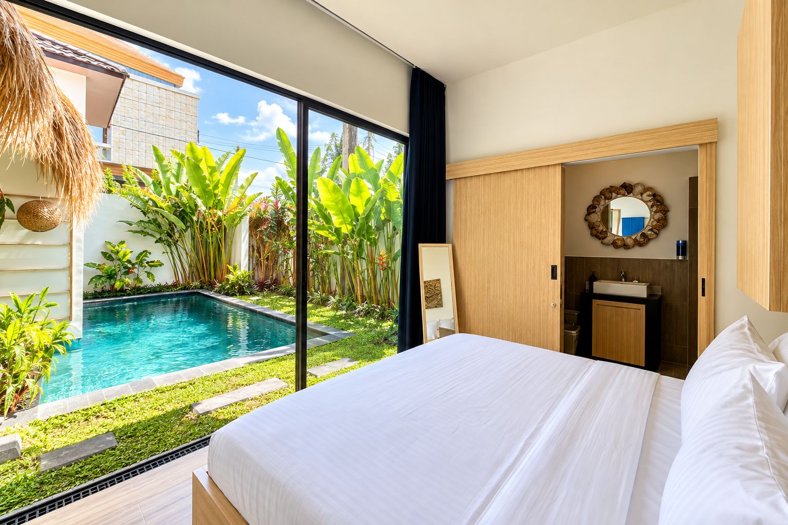 A bedroom with a large bed, a bathroom with a sink, and a view of a swimming pool with tropical plants.