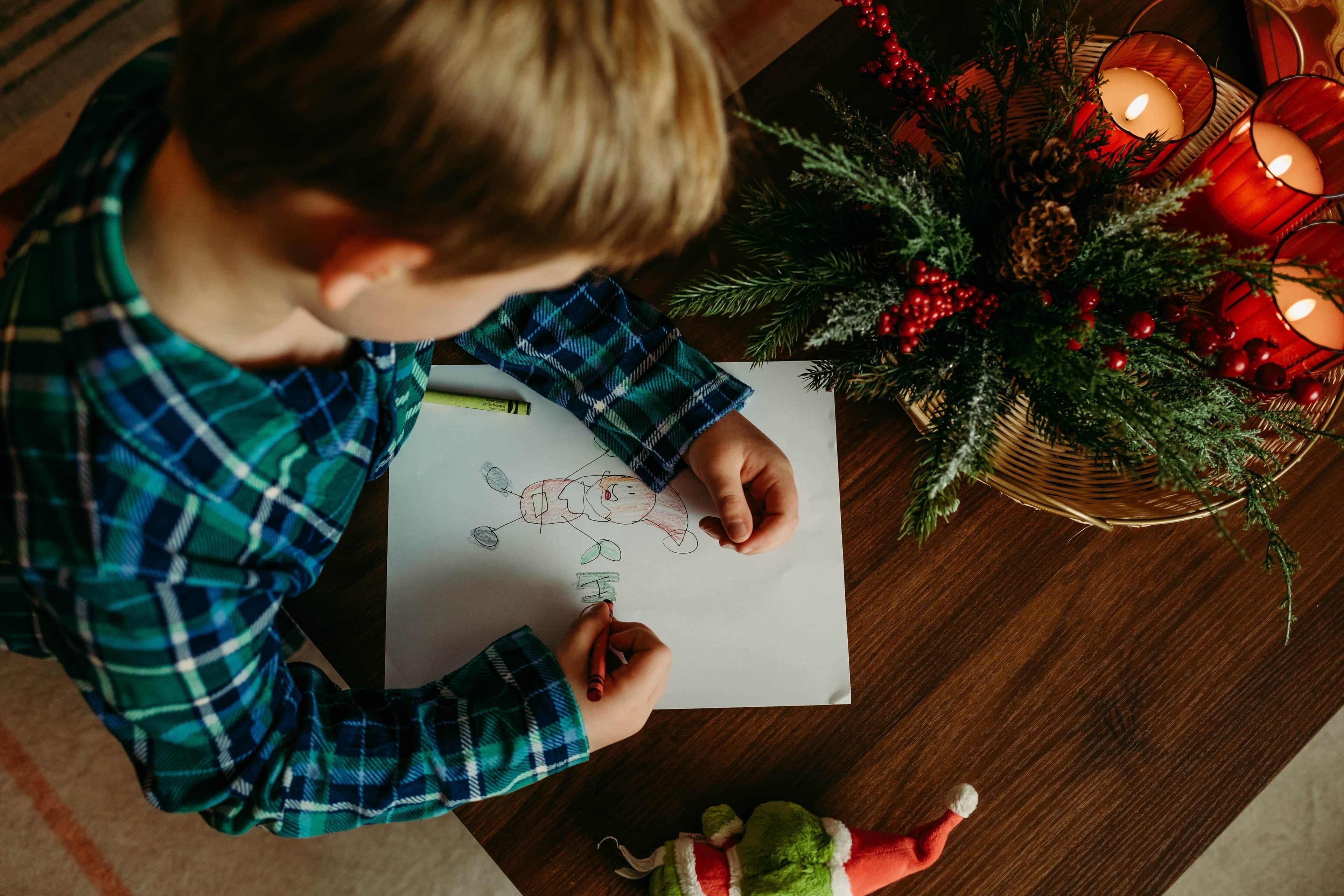 A child drawing on paper at a wooden table with a Christmas decoration and candles.
