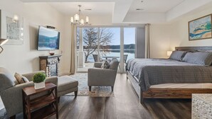 A modern bedroom with a large bed, a sofa, a small table, and a TV. There is a view of a lake through the glass door. - Water`s Edge Luxury Retreat at Delton Grand (Wisconsin Dells)
