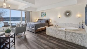 A bedroom with a large bed, a sitting area with a table and chairs, a bathroom with a bathtub, and a view of a lake through the window. - Water`s Edge Luxury Retreat at Delton Grand (Wisconsin Dells)