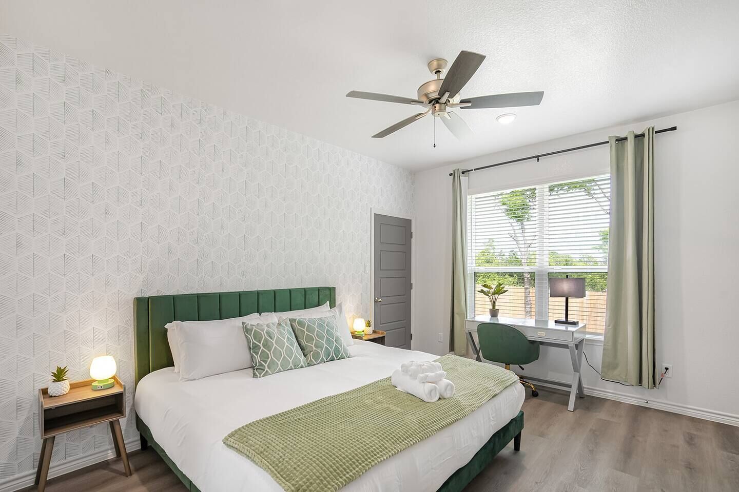 A hotel room with a bed, a desk, a chair, a ceiling fan, and a window with curtains.
