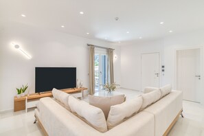 A modern living room with a flat-screen TV, a sofa, a coffee table, and a large window with curtains. - Villa Raphael - Luxury and Serenity in Paros (Naousa)