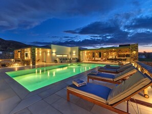 A modern outdoor pool area with lounge chairs and a stone wall. - Villa Raphael - Luxury and Serenity in Paros (Naousa)