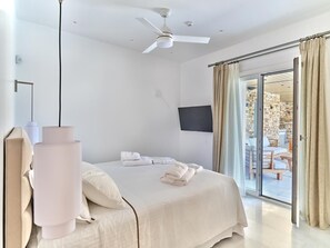 A modern hotel room with a bed, a ceiling fan, a TV, and a sliding glass door to an outdoor area. - Villa Raphael - Luxury and Serenity in Paros (Naousa)