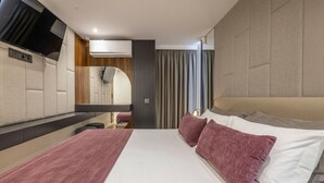 Ein modernes Hotelzimmer mit einem großen Bett, einem wandmontierten Flachbildfernseher, einem Schminktisch mit Spiegel und einem großen Fenster mit Vorhängen.