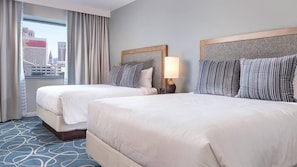 Ett hotellrum med två sängar, utsikt över staden, ett stort fönster med gardiner och en sänggavel med en strukturerad panel.