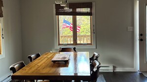 Une salle à manger avec une grande table en bois, six chaises, un luminaire suspendu et une fenêtre ornée d’un drapeau américain.