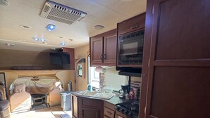A compact RV kitchen with a microwave, sink, and stove, featuring wooden cabinets and a small rug. - Cozy RV stay on a South Gilbert Mini Farm (Gilbert)