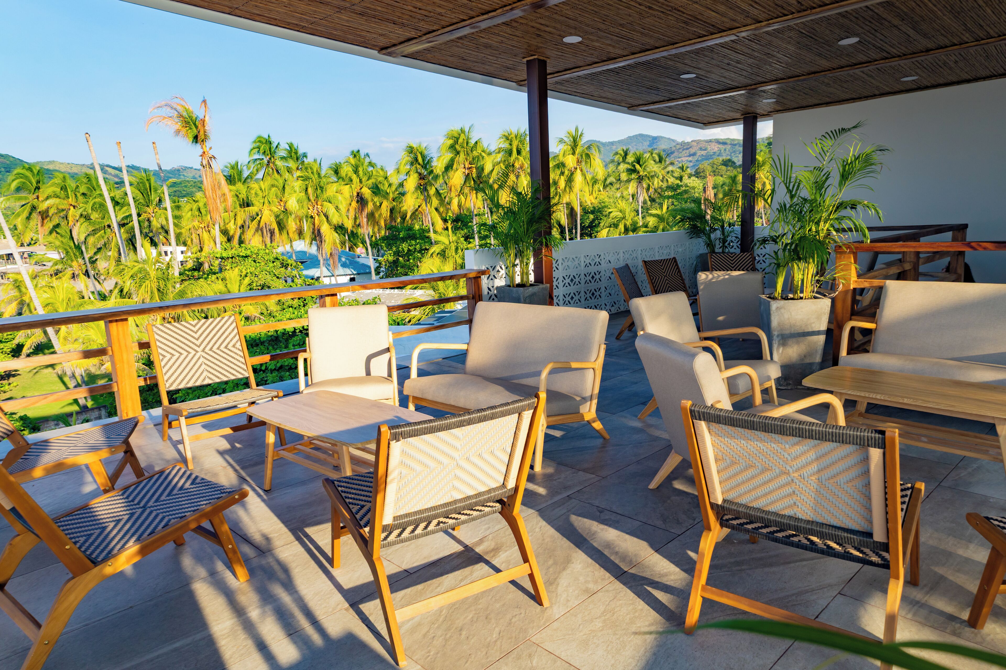 A covered outdoor seating area with wicker furniture, a wooden table, and a view of palm trees and mountains.