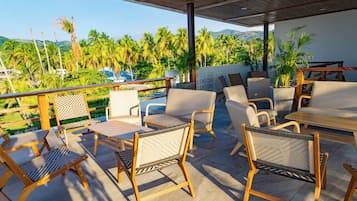 A covered outdoor seating area with wicker furniture, a wooden table, and a view of palm trees and mountains.