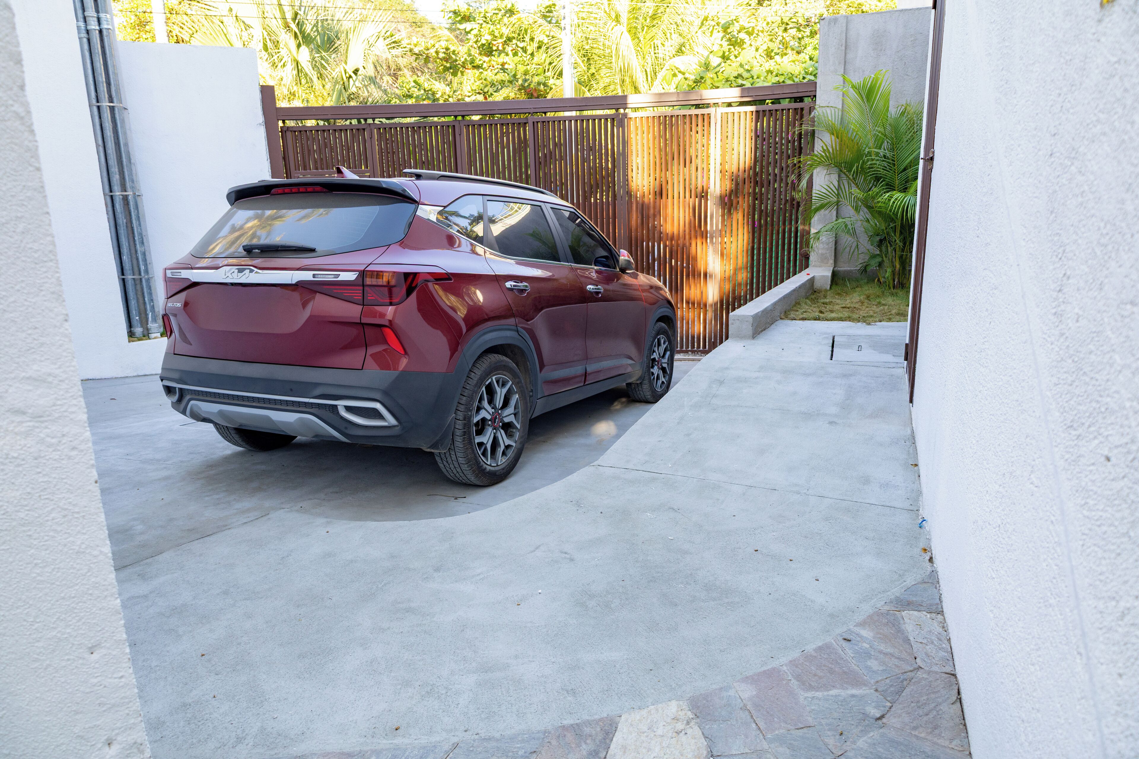 A maroon SUV parked in a covered driveway with a stone-tiled path, a wooden fence, and a white wall.