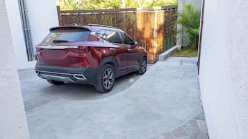 A maroon SUV parked in a covered driveway with a stone-tiled path, a wooden fence, and a white wall.