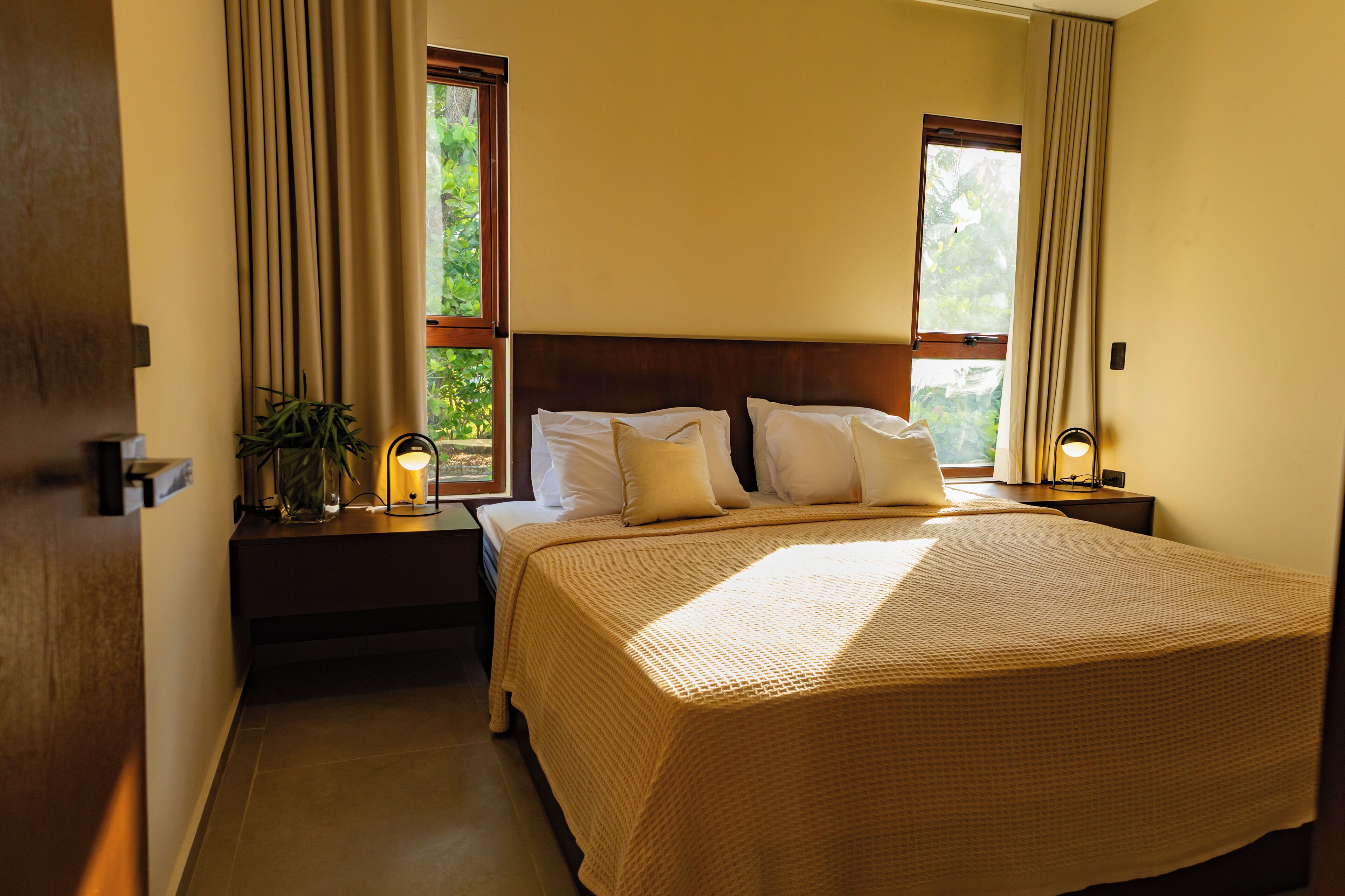 A neatly made bed with a beige bedspread and pillows, two bedside lamps, a nightstand with a plant, and a window with curtains.