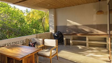 A patio with a wooden table and chairs, a grill, and a view of lush greenery.